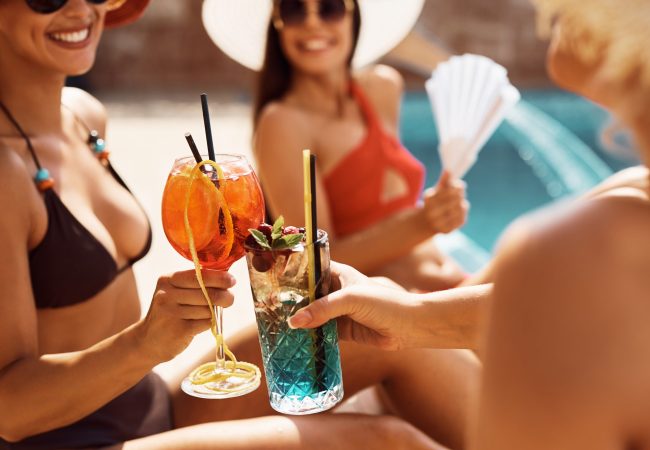 Close up of female friends toasting while drinking cocktails at swimming pool during summer day.